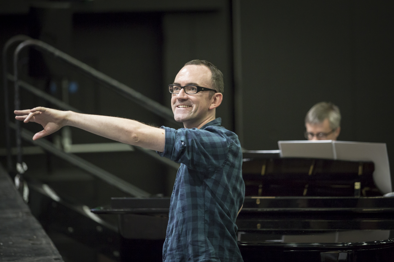 Image. A man with short brown hair and glasses, wearing a blue plaid shirt. He is smiling and extending
his arms expressively as if conducting or directing, with a grand piano in the background.