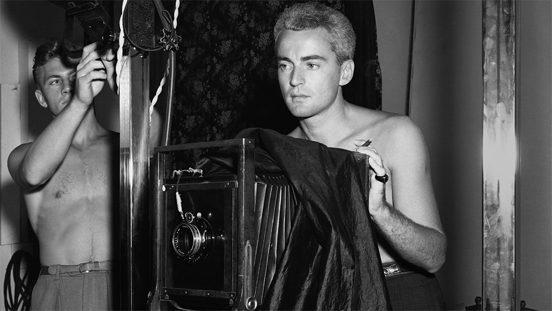 Black and white photo of two white men without shirts on. One is standing behind a large box camera, possibly from the 1940s. The second man is helping to set up equipment in the background.