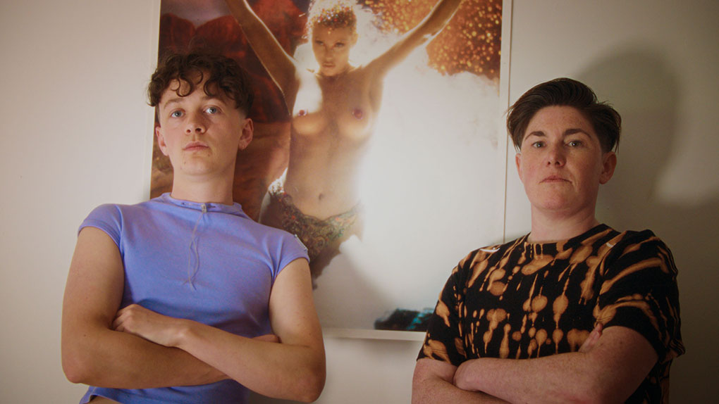 Two young white people with their arms folded, standing in front of a framed picture of a topless woman.