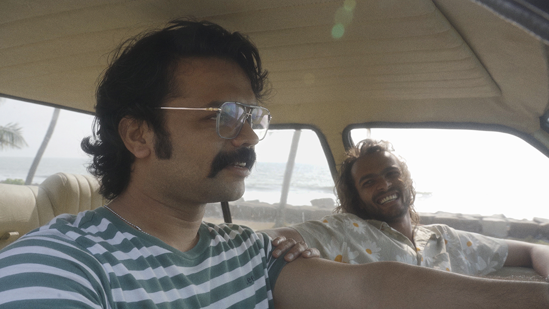 Two men are sitting in a car, with one man in the driver's seat wearing glasses and a striped shirt, and the other man in the passenger seat, smiling and wearing a light-coloured shirt with a floral pattern. The ocean and palm trees are visible through the windows.