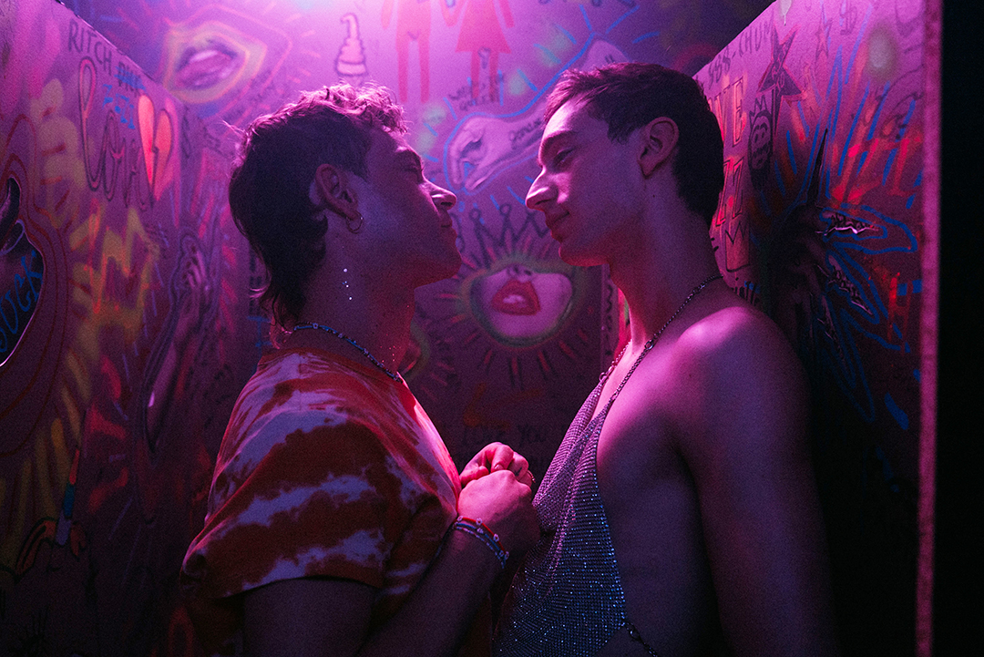 Two people standing in what could be a graffiti covered bathroom of a nightclub. They are staring into each other's eyes. The person on the left is wearing a red and white tie-dye t-shirt. The person on the right is wearing a shimmering silver tank top or dress..